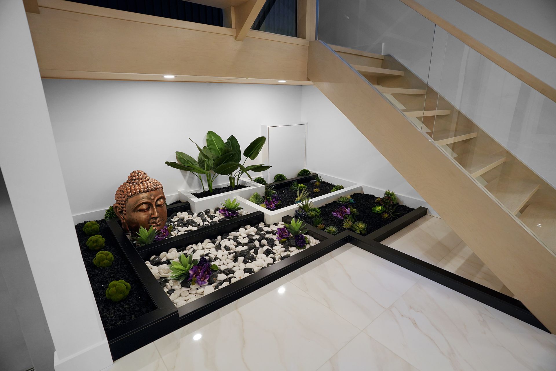 A modern zen garden with a Buddha head, plants, and white and black stones, under a staircase.