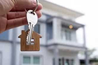 Hand holding house key with home-shaped keychain in front of a modern house.