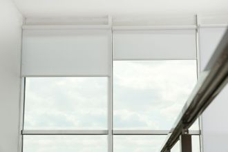 Two windows with white roller shades, partially lowered, against a cloudy sky.