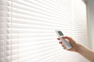Person's hand holding a remote, controlling white horizontal blinds. The blinds are partially open in a bright room.