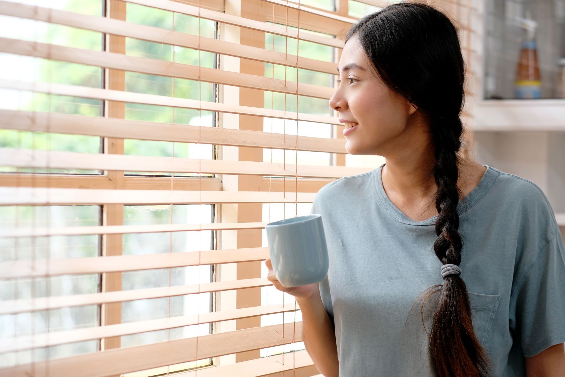 Woman with braid holding mug, looking out a window with wooden blinds.