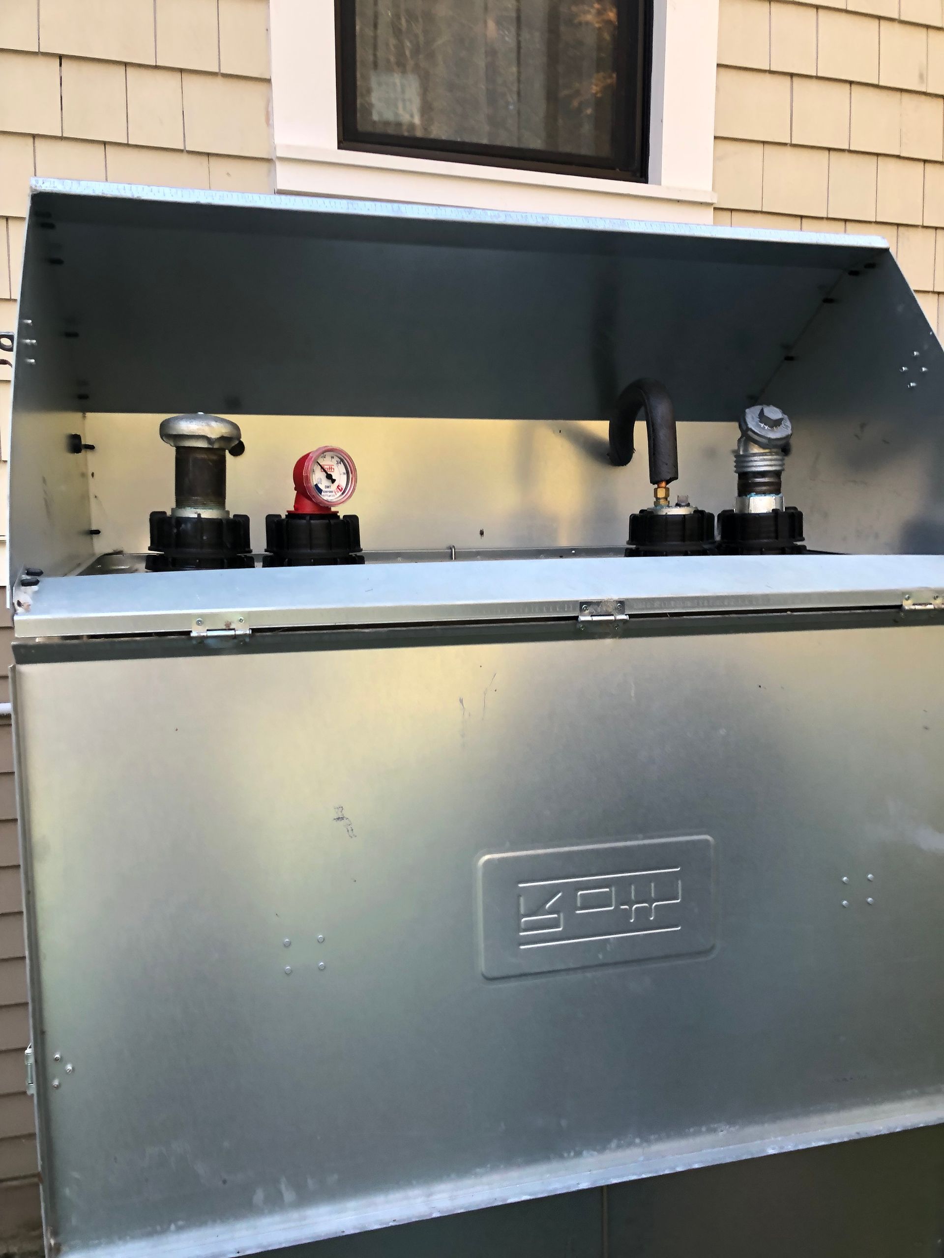 Outdoor metal utility box with valves and pipes mounted on top, under a window.