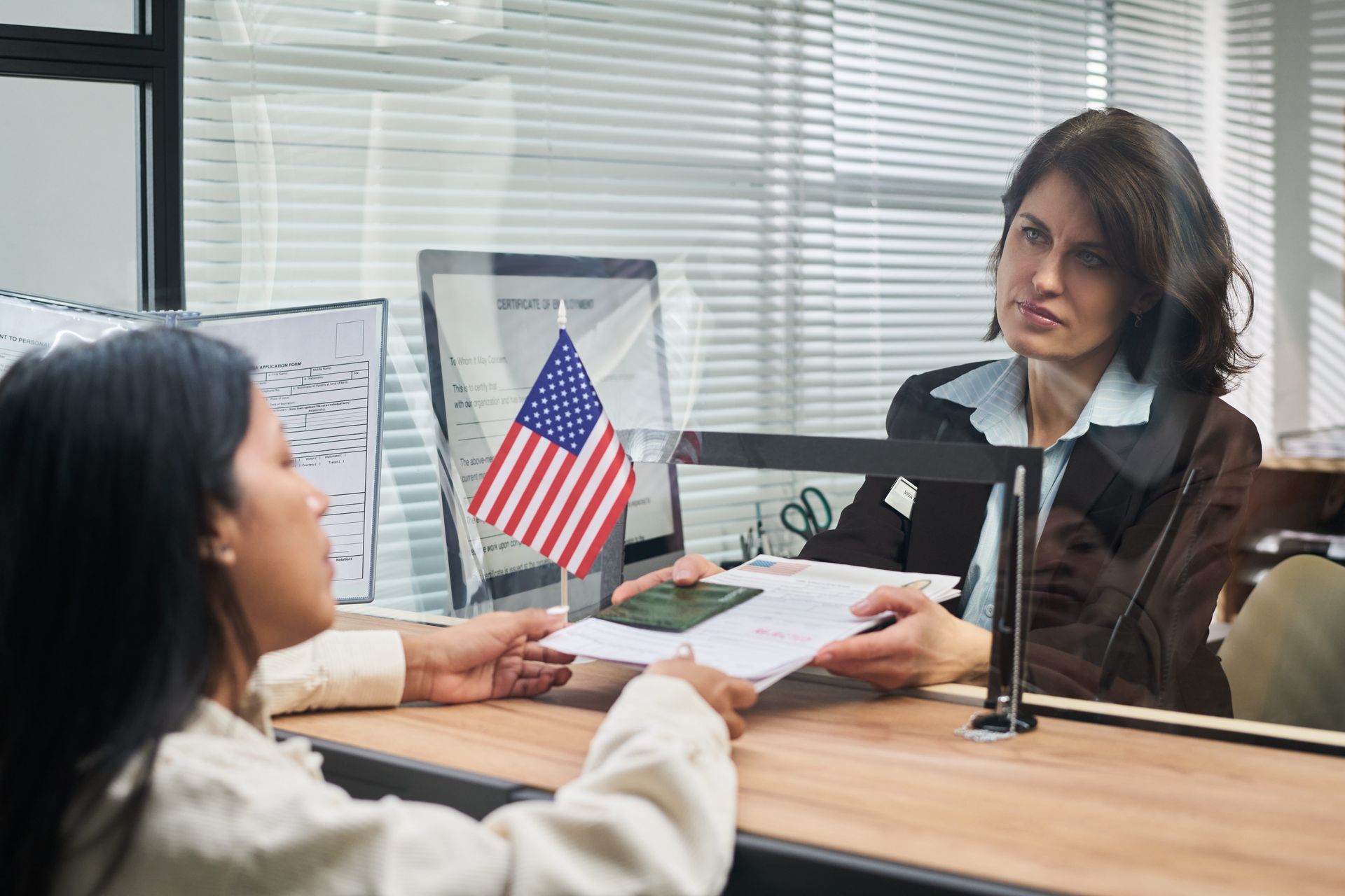 Two women at a desk across a glass partition, with a small U.S. flag between them.