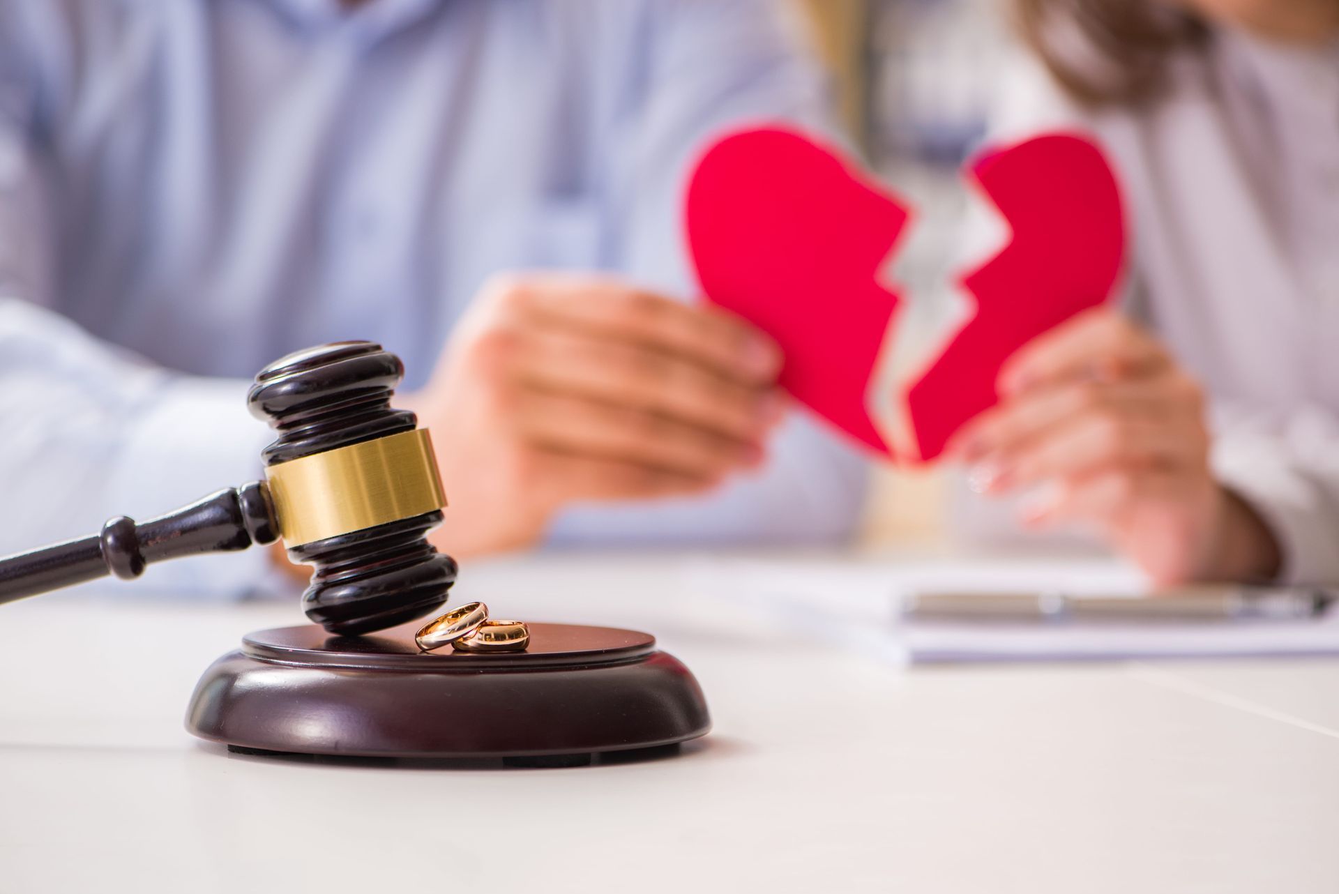 A judge's gavel rests on a sound block with two rings beside it, as two people in the background hold a broken red heart.