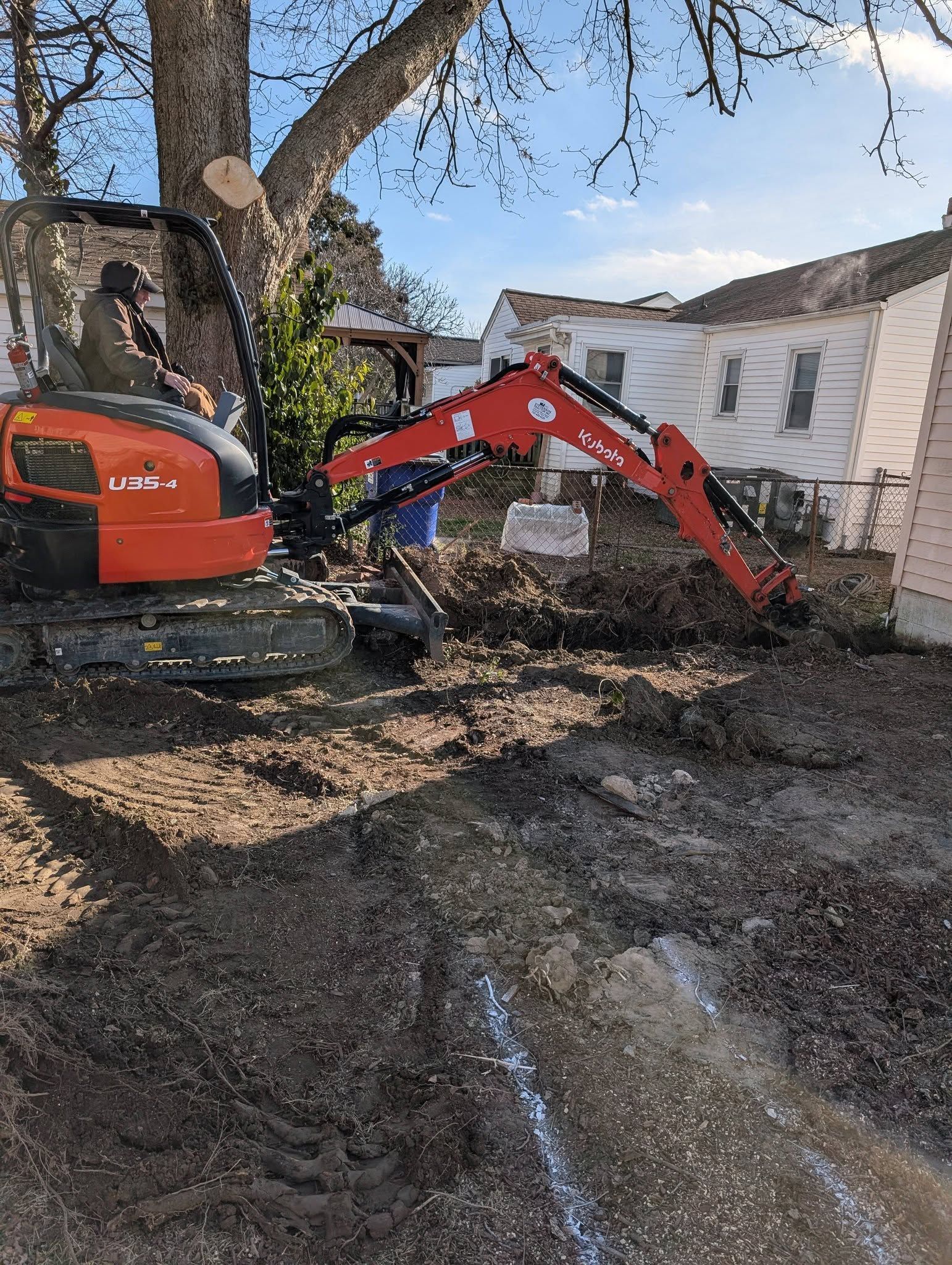 Orange excavator digging near a white house in a muddy yard beside a large tree