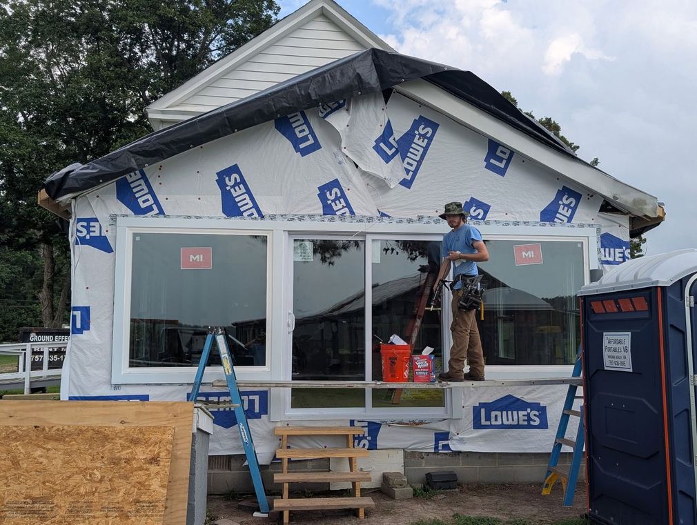 House under construction with workers on a porch, wrapped in white house wrap and window openings covered.