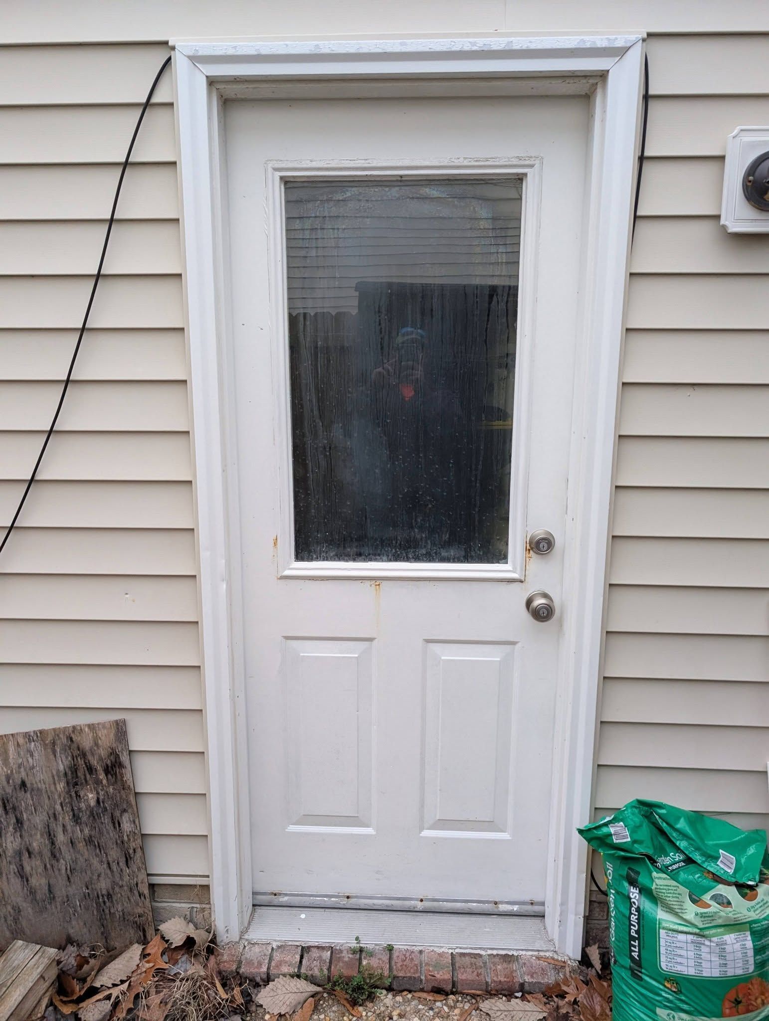 White exterior door with a window in beige siding; a green bag sits by the lower right corner.