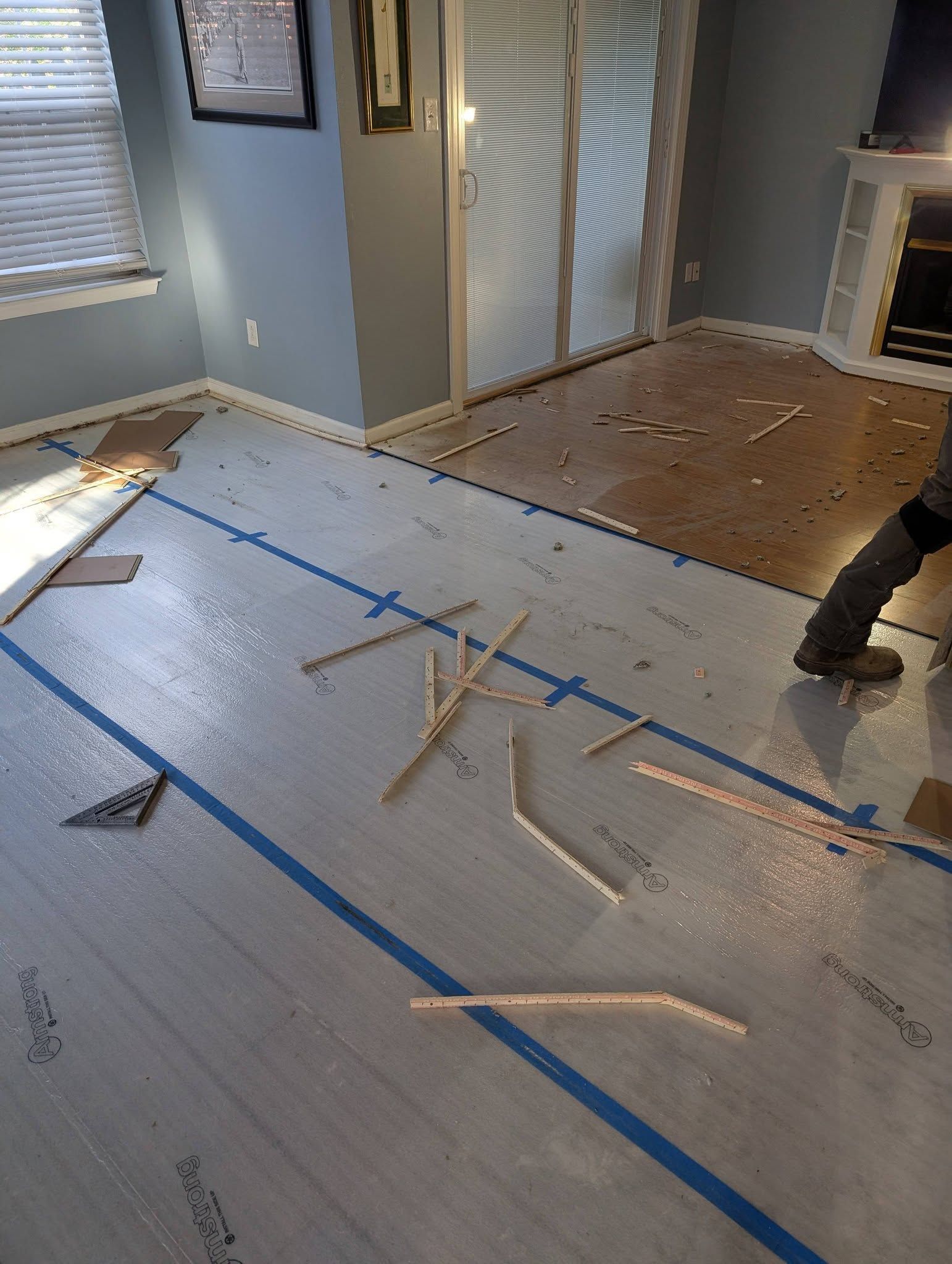 Living room floor under renovation with underlayment, blue tape lines, and a worker’s legs visible