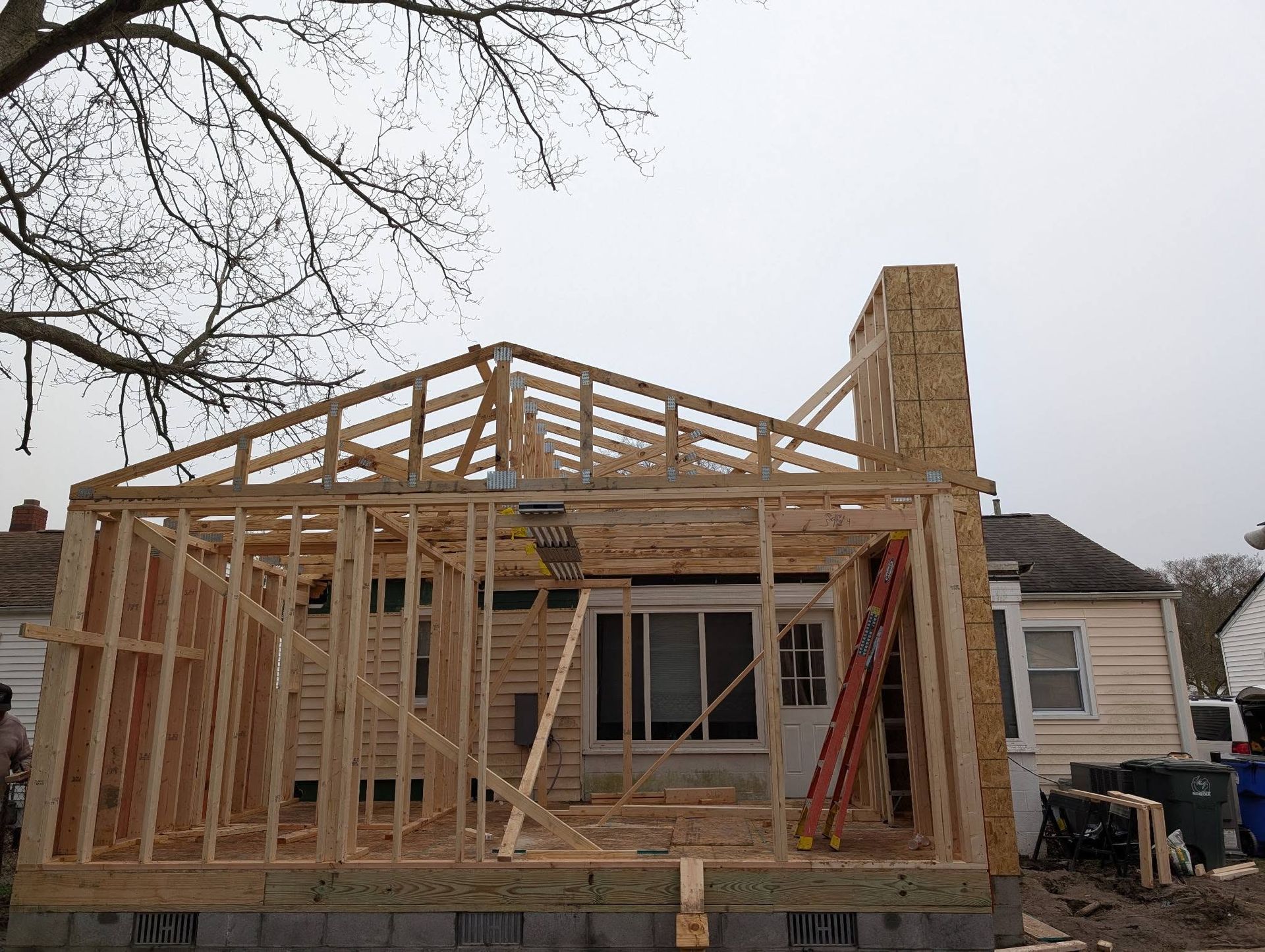 Wood-framed house under construction with roof trusses and a tall stone chimney under an overcast sky