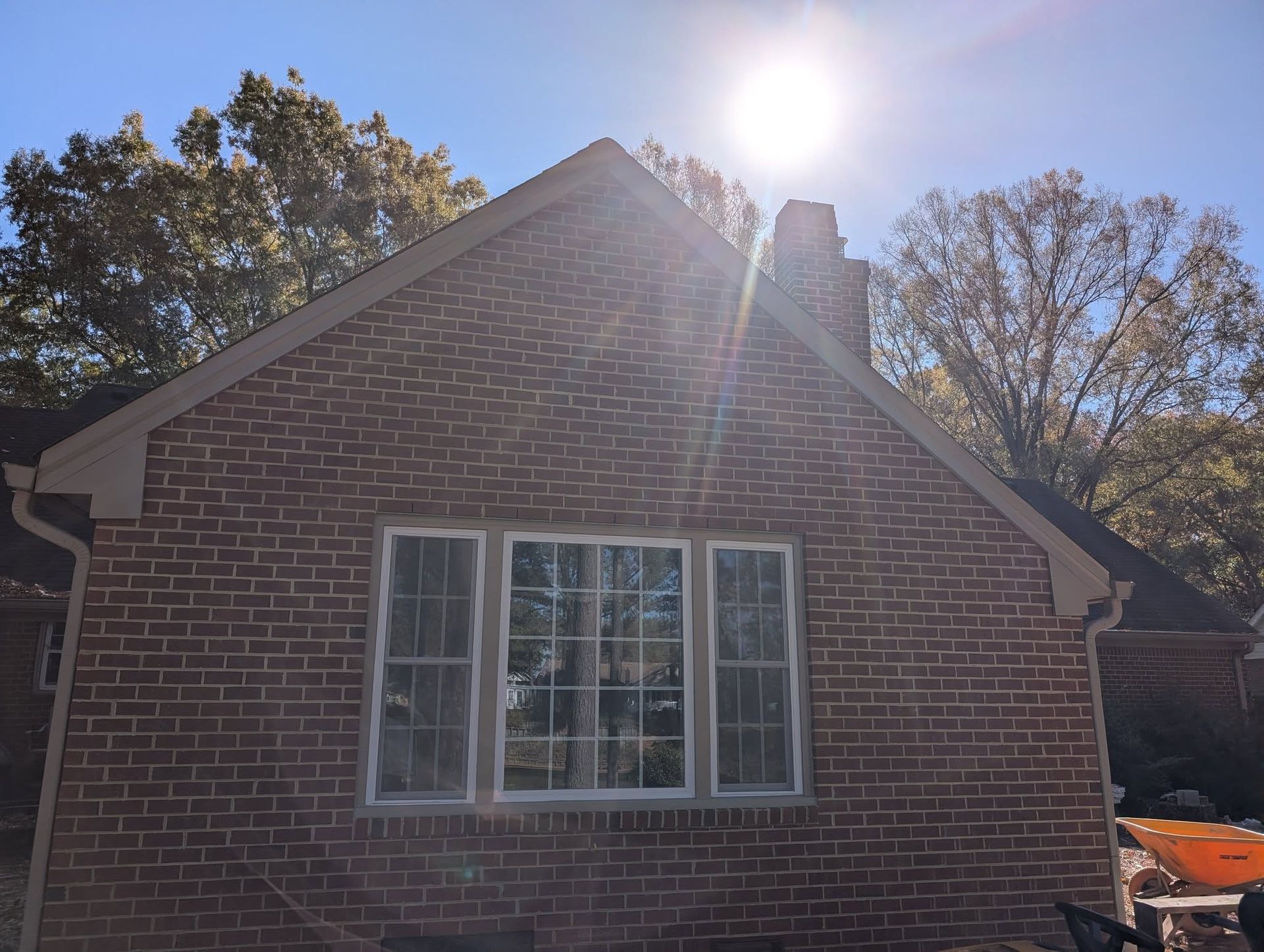 Brick house gable with white-trimmed window under bright sun, trees in the background