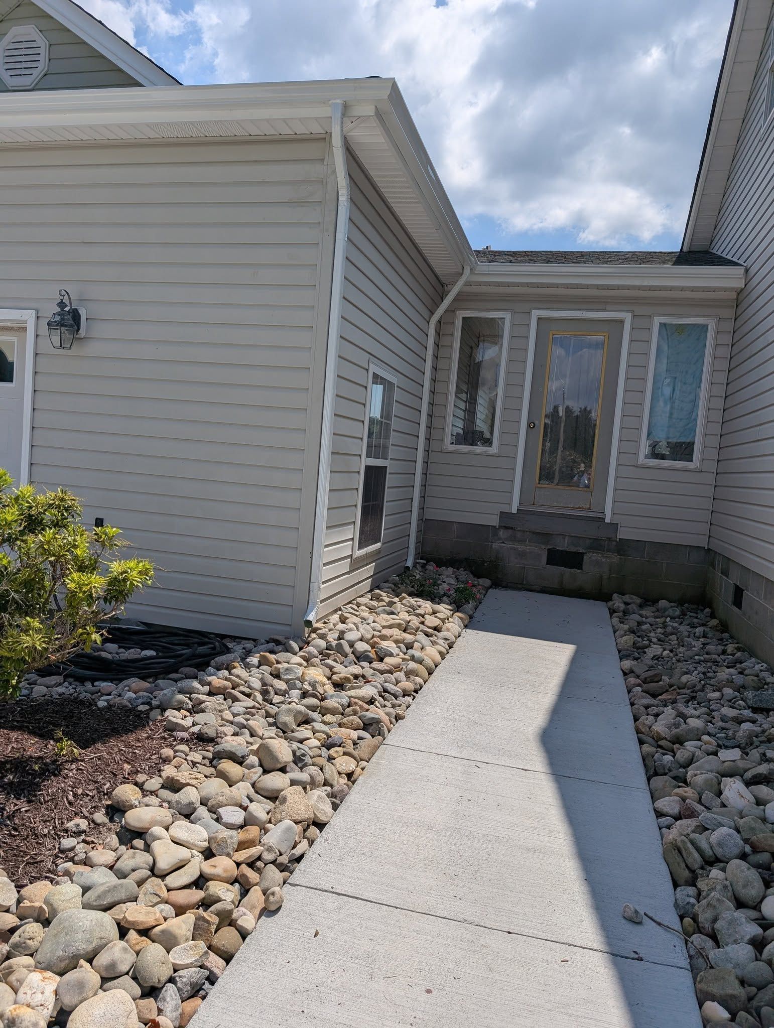 Concrete walkway leading to a front door between a house wall and rock landscaping under a cloudy sky