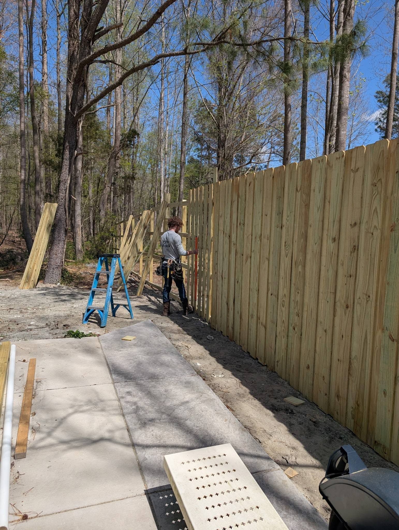Worker standing by a tall wooden fence on a concrete path beside a blue ladder in a wooded yard