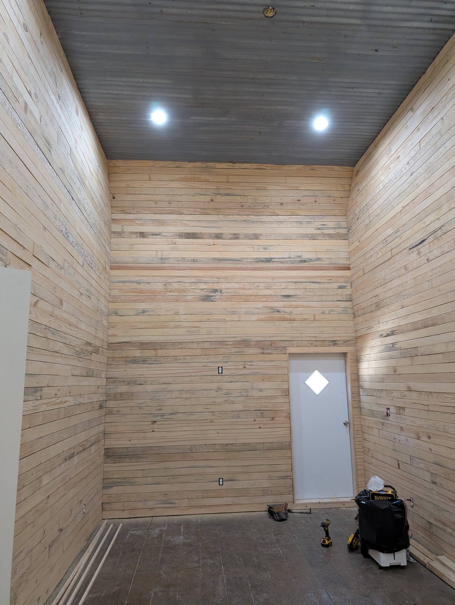Empty high-ceiling room with wood-paneled walls, two ceiling lights, and a white door on the back wall.