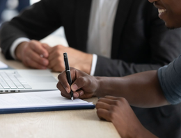 Hands signing a document; a small house model sits nearby on a wooden table.