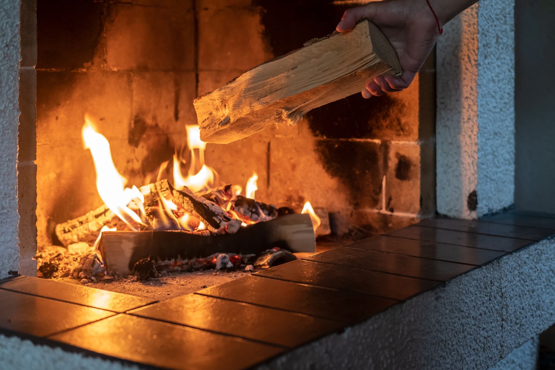 A hand placing firewood into a fireplace with flames.