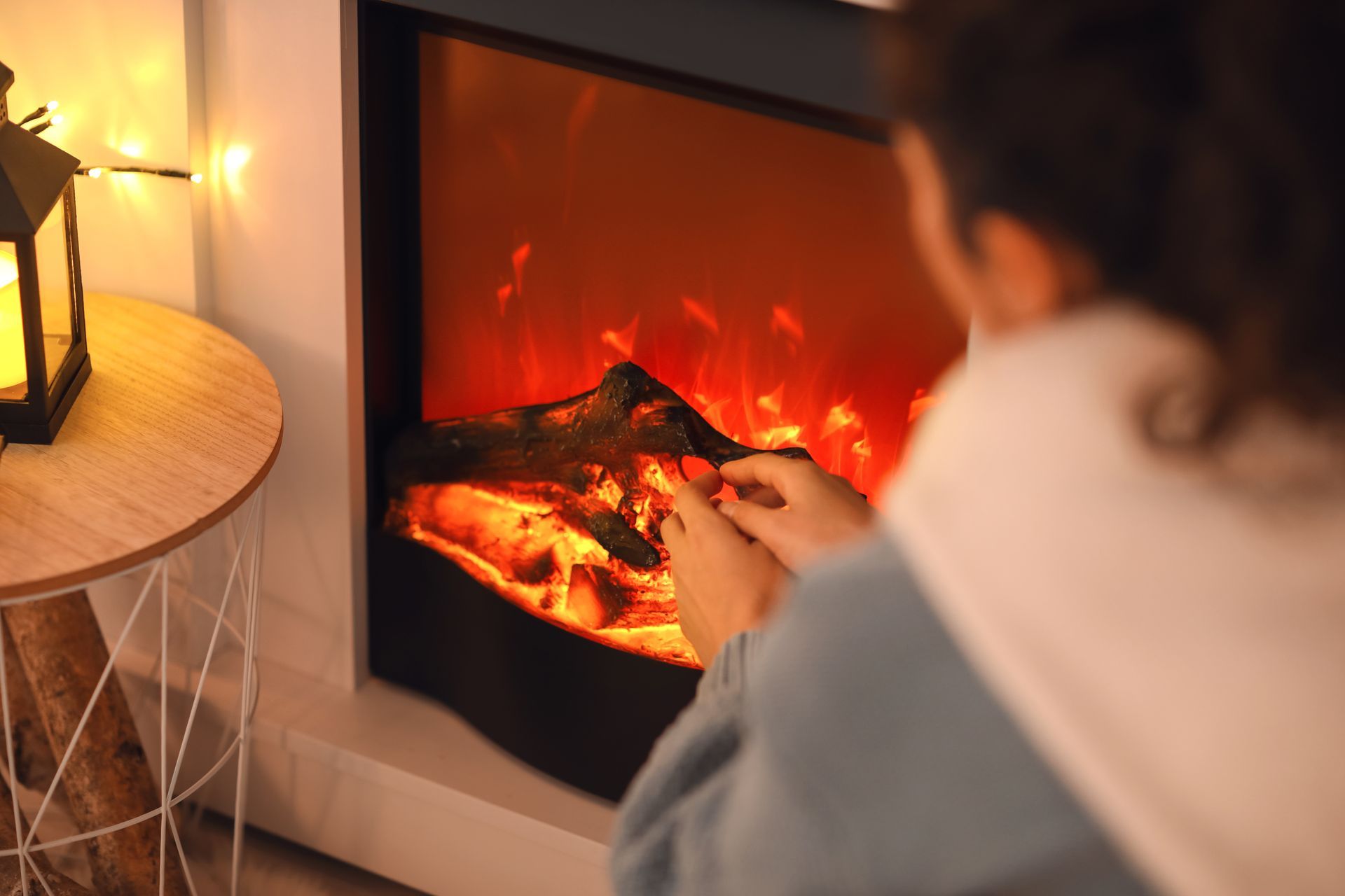 Person warming hands near a glowing electric fireplace; a lantern and side table are visible.