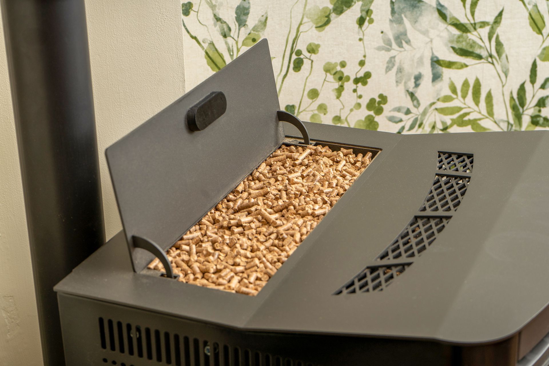 Pellet stove with open lid revealing wood pellets, with a black flue pipe and floral wallpaper in the background.