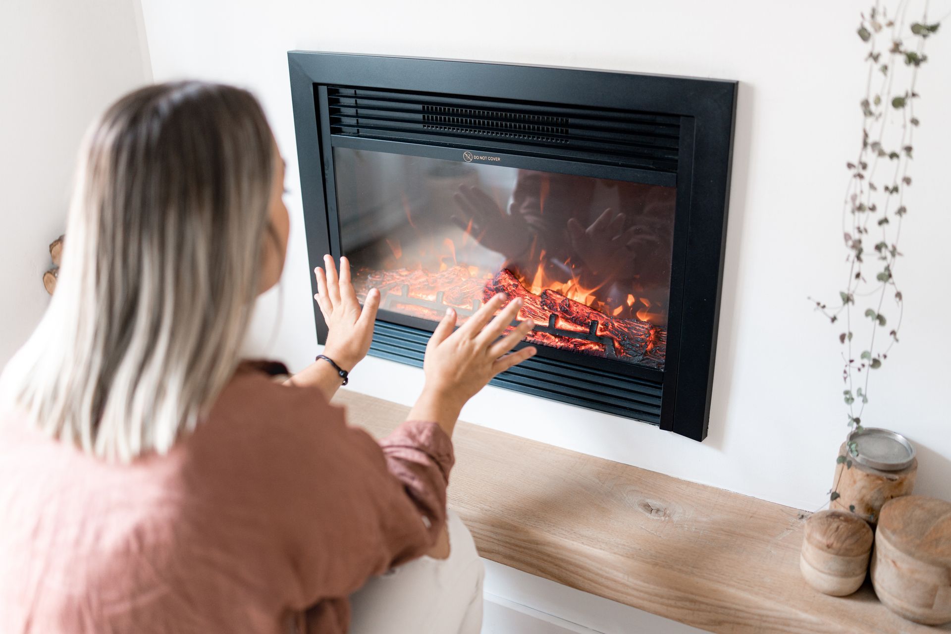 Woman warms hands near a black electric fireplace, set in white wall.