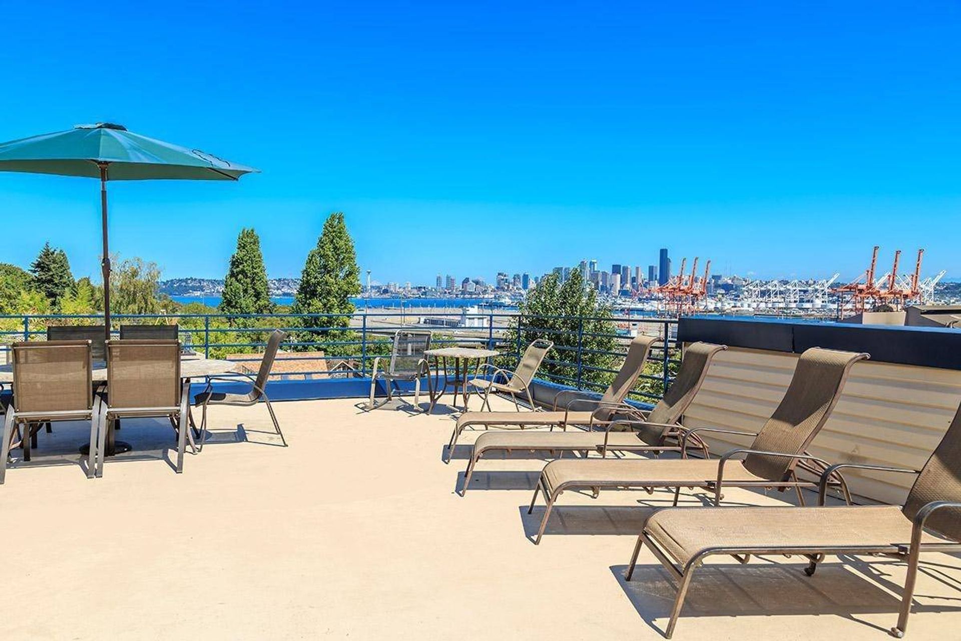 Sunny rooftop terrace with lounge chairs and umbrellas overlooking a city skyline and waterfront.