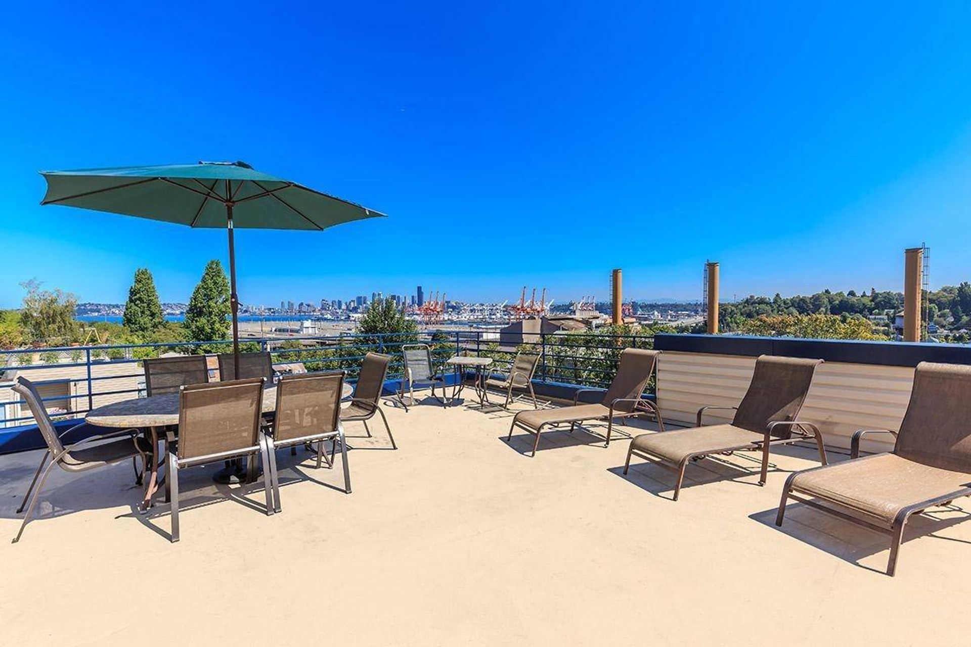 Rooftop patio with lounge chairs, dining table, green umbrella, and city skyline under a bright blue sky
