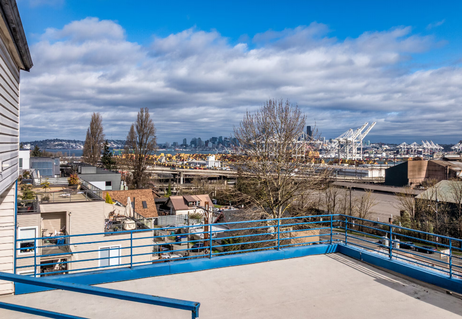 Rooftop terrace with blue railings overlooking a harbor, city buildings, and cloudy sky