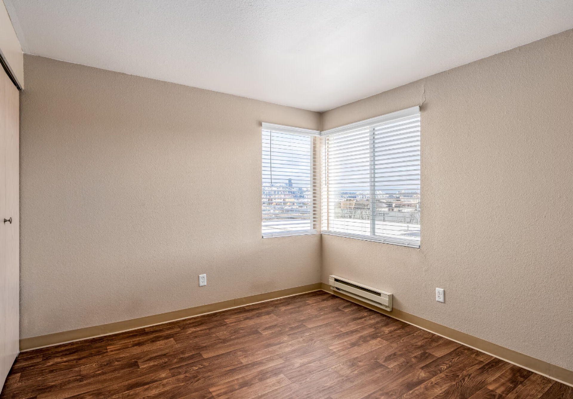Empty beige room with wood floor, corner windows, and white blinds