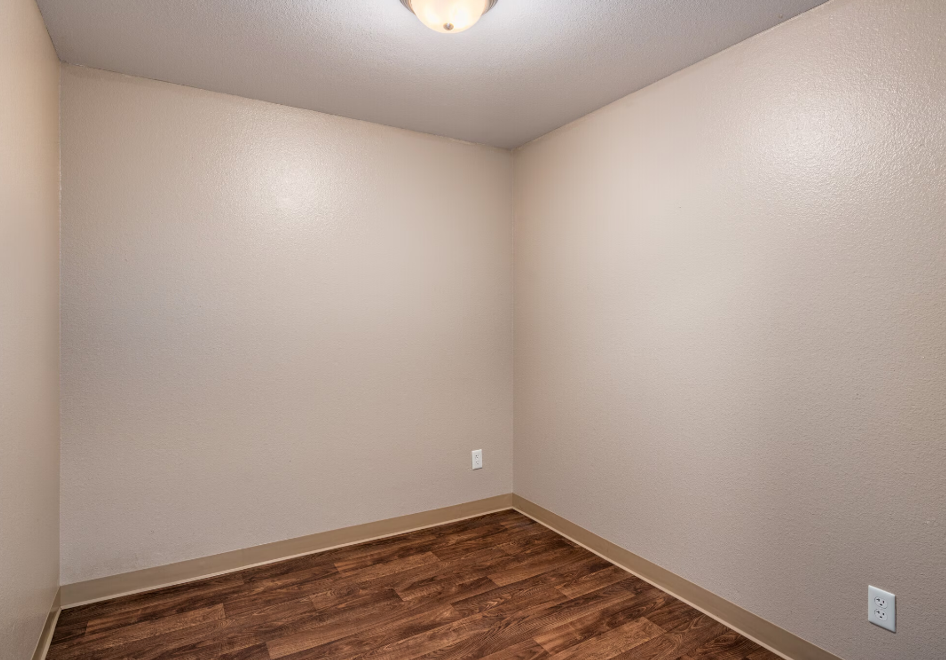 Empty beige corner room with wood-look floor and white electrical outlets