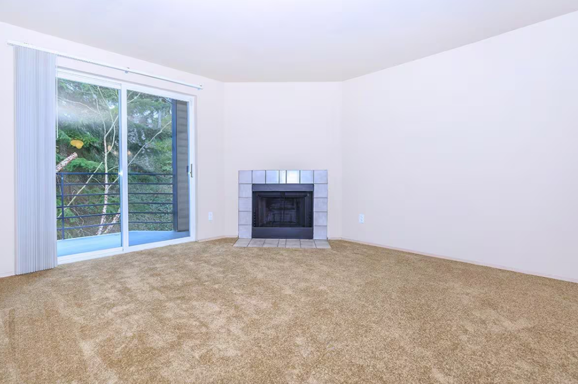 Empty beige-carpeted living room with white walls, a fireplace, and sliding glass doors to a patio.