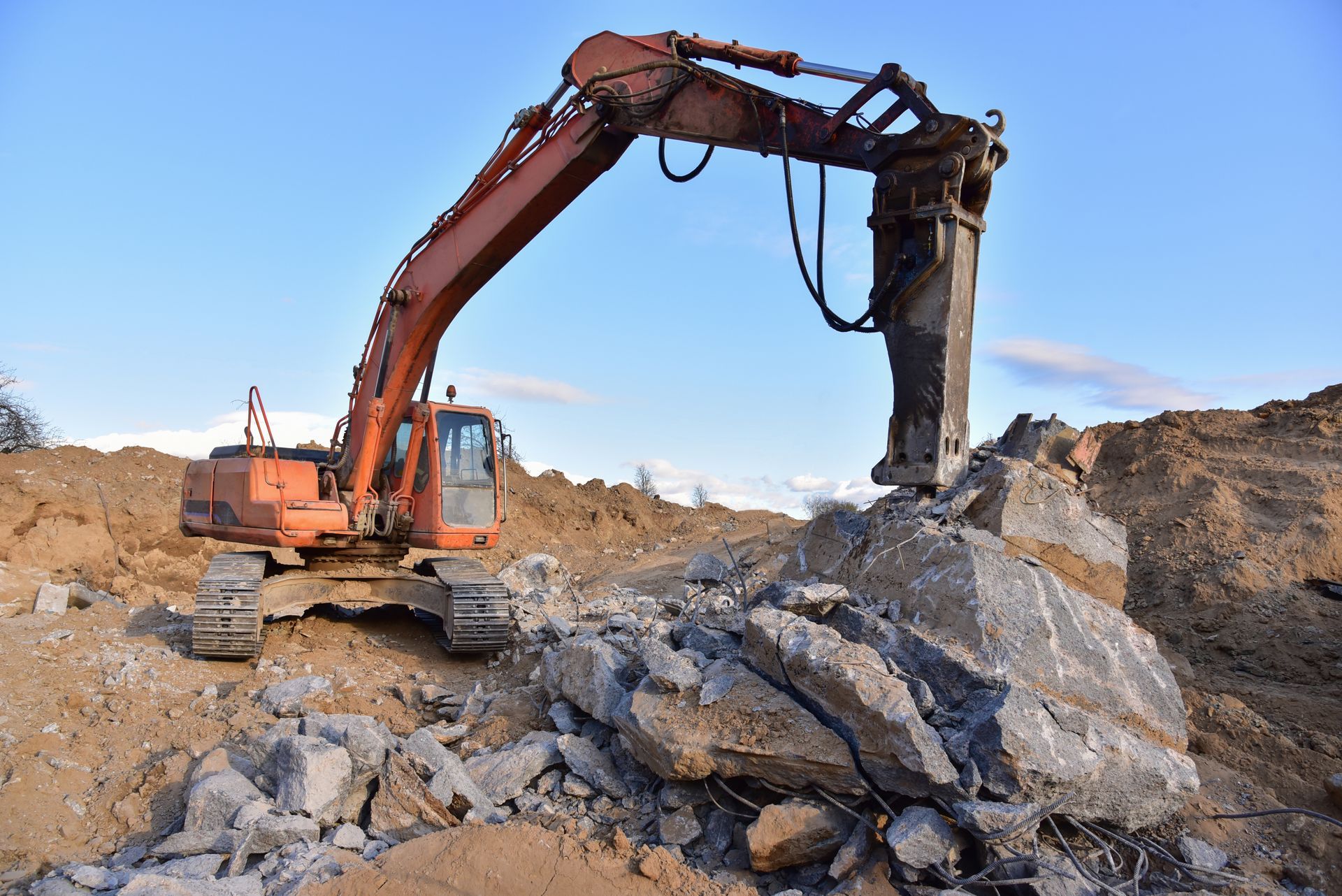 Orange excavator using a hydraulic hammer to break apart a large concrete block at a construction site.