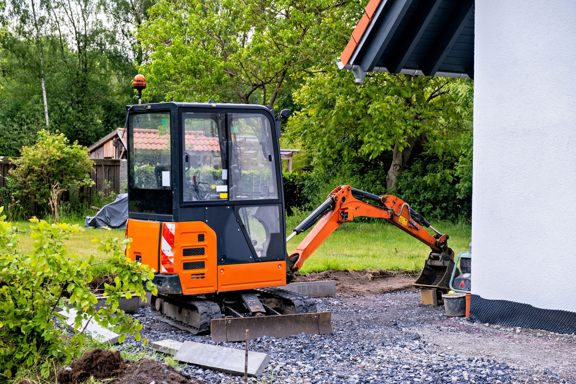 Orange and black mini excavator digging next to a white building. Green grass and trees in the background.