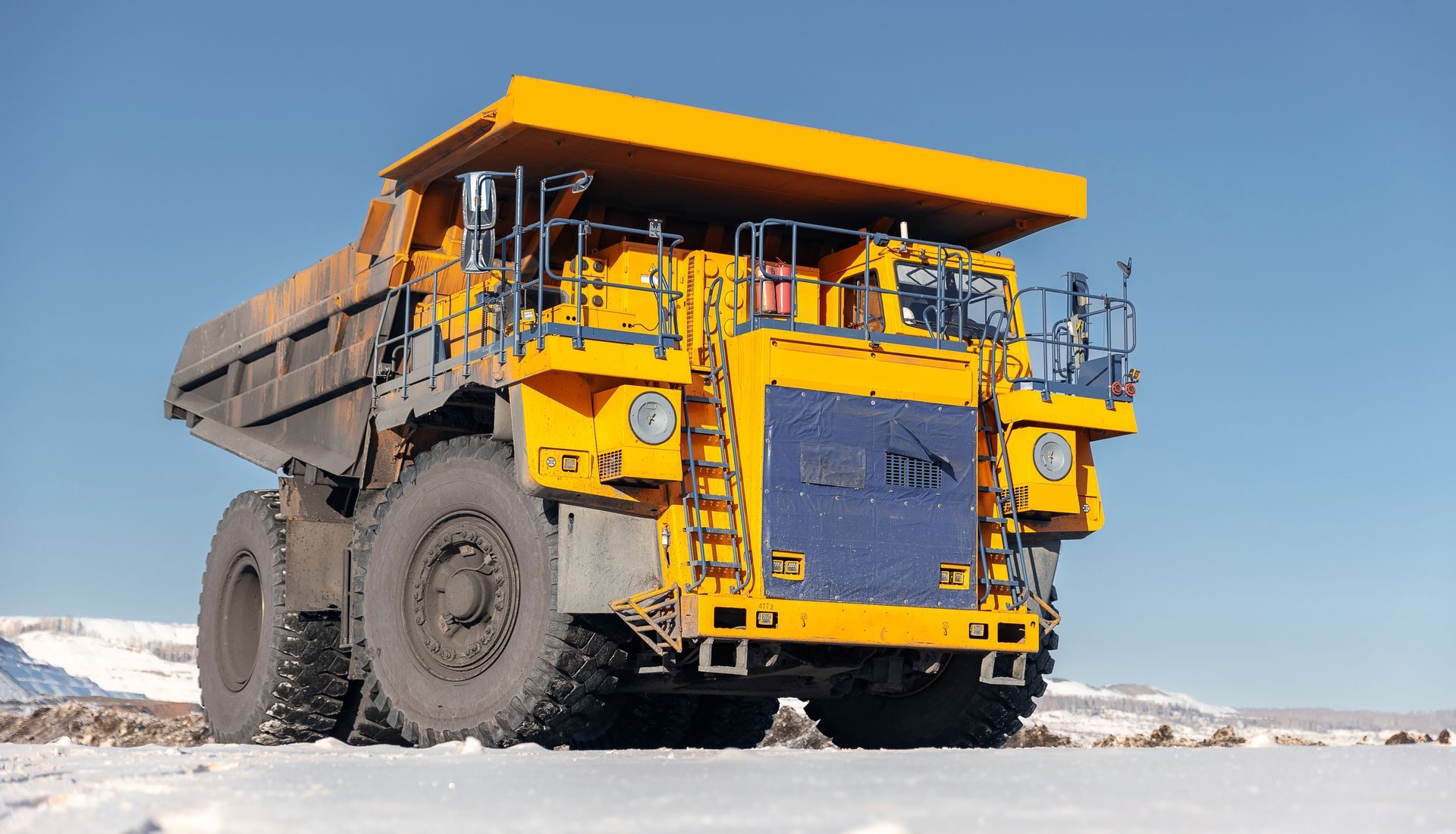 Yellow mining dump truck on snow-covered ground against a clear blue sky.