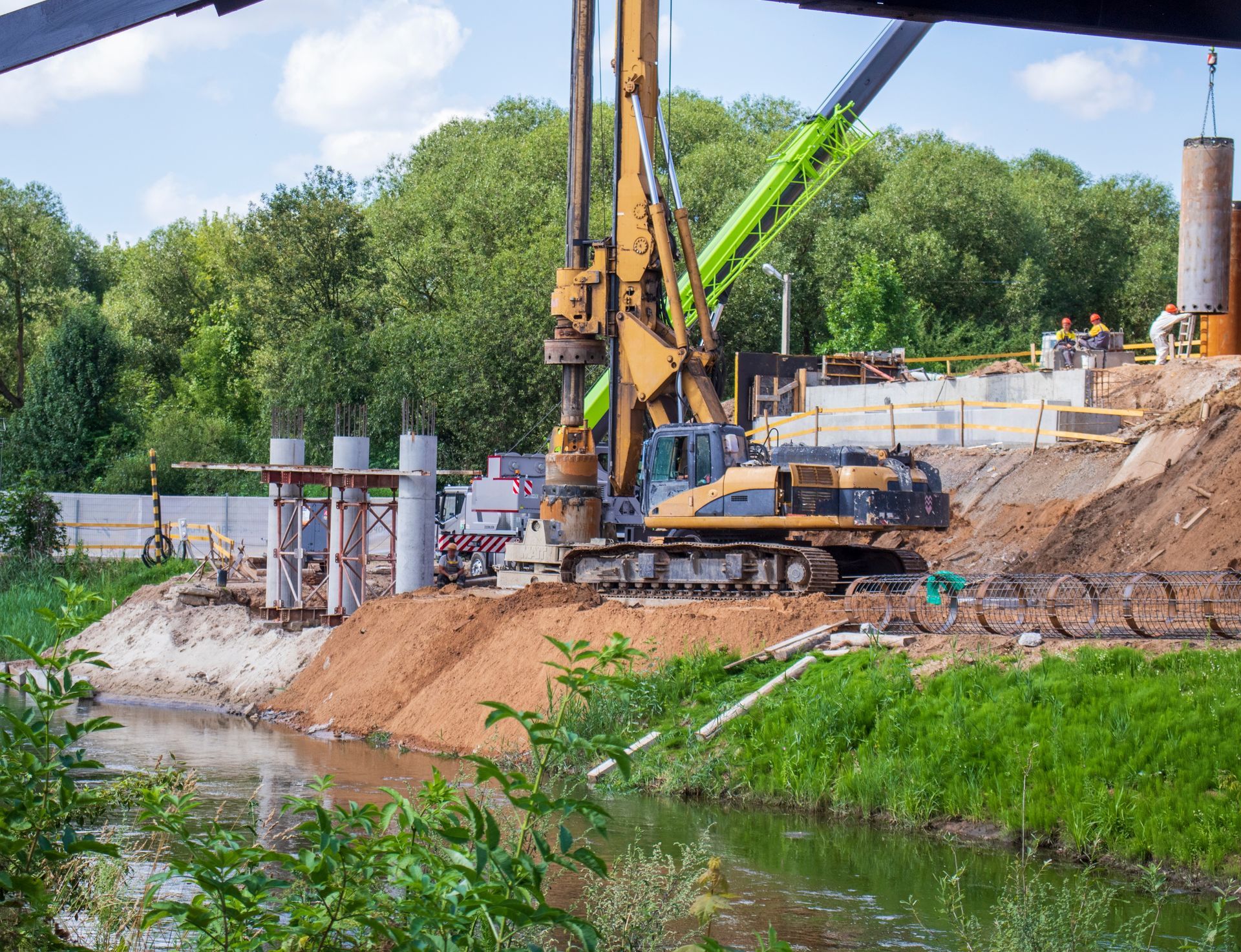 Construction site: excavator drilling, workers, concrete pillars near a river.