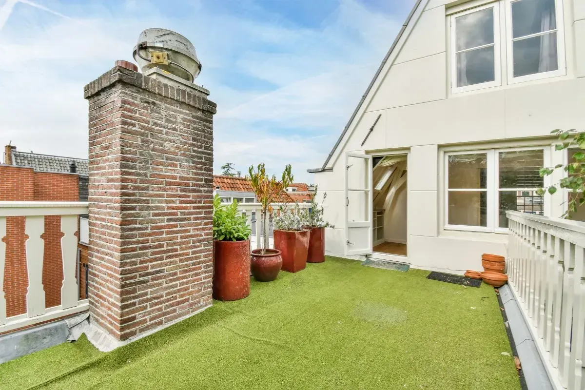 Henderson Artificial Turf- A rooftop terrace with artificial grass, red brick chimney, and potted plants. White building facade with windows and a clear sky overhead. Cozy and inviting.