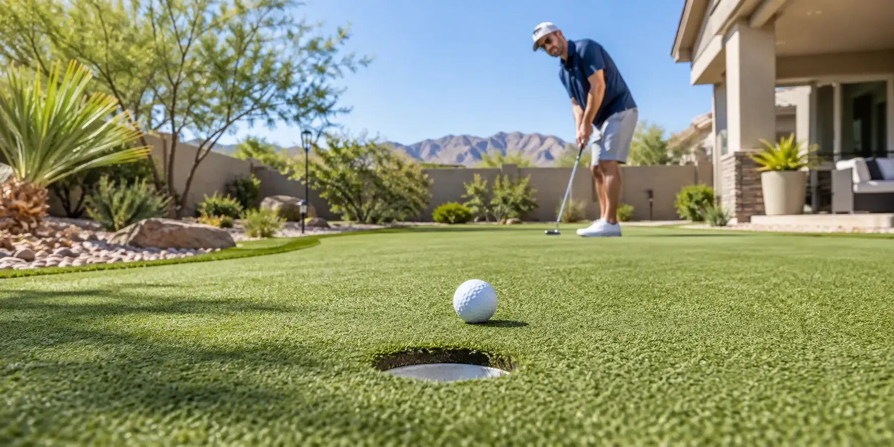 A golf ball on a pristine artificial putting green in a sunny backyard with a golfer practicing in the background.
