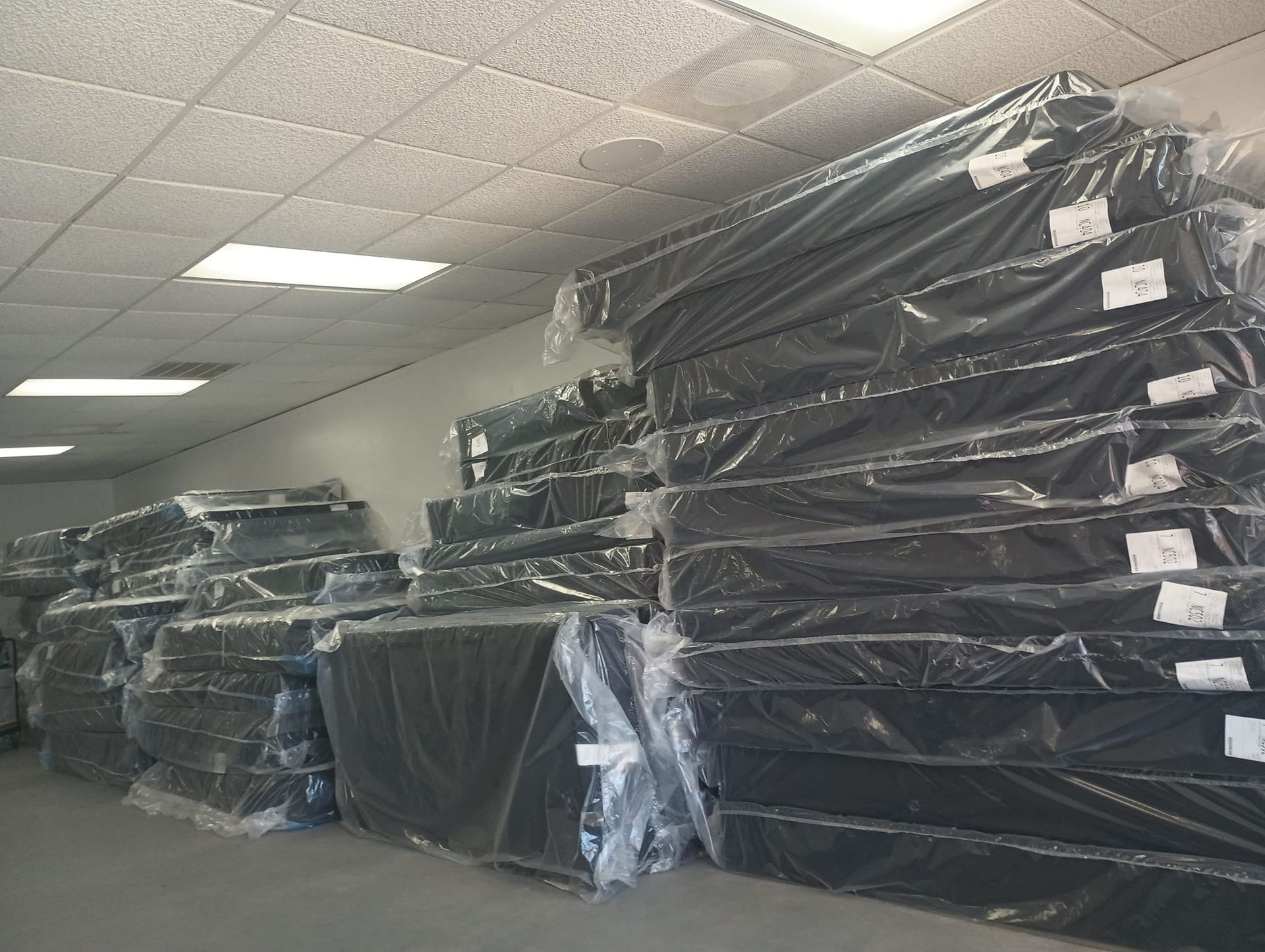 Stacks of black mattresses wrapped in plastic, stored in a room with fluorescent lighting.