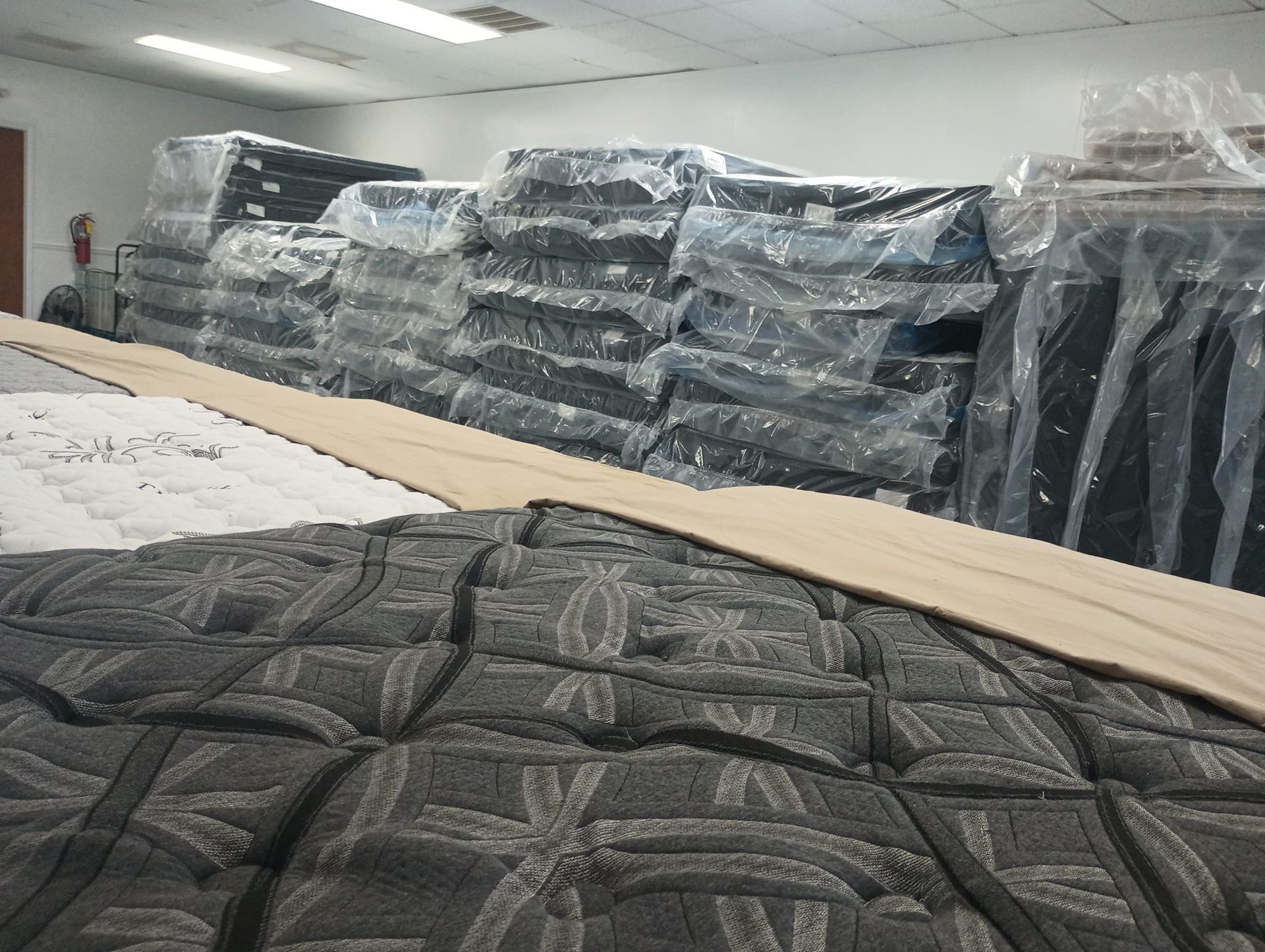 Mattresses in a storage room, some wrapped in plastic. Focus on textured patterned grey and tan bedding.