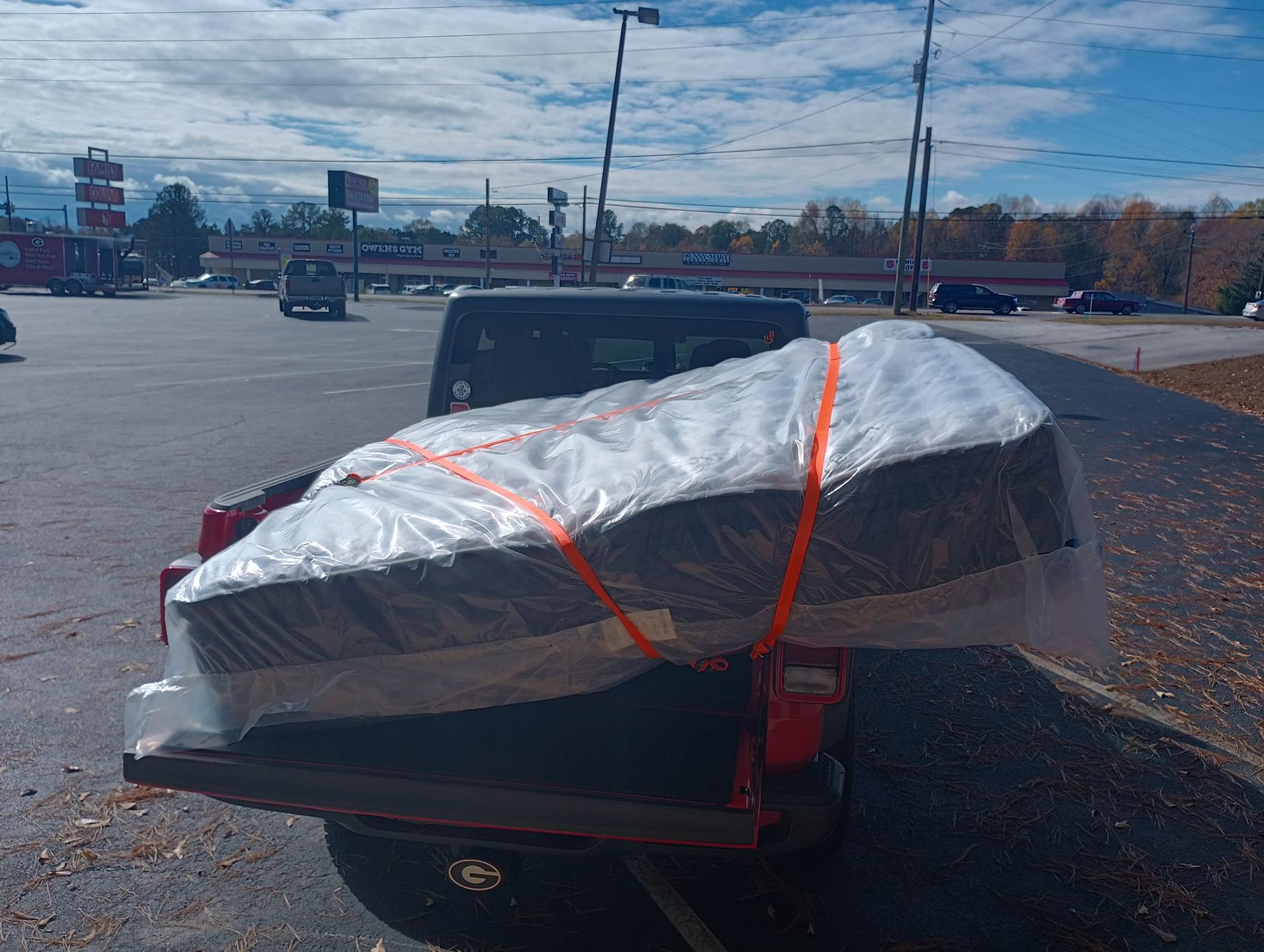 Red pickup truck bed carrying a mattress wrapped in plastic and secured with orange straps in a parking lot.