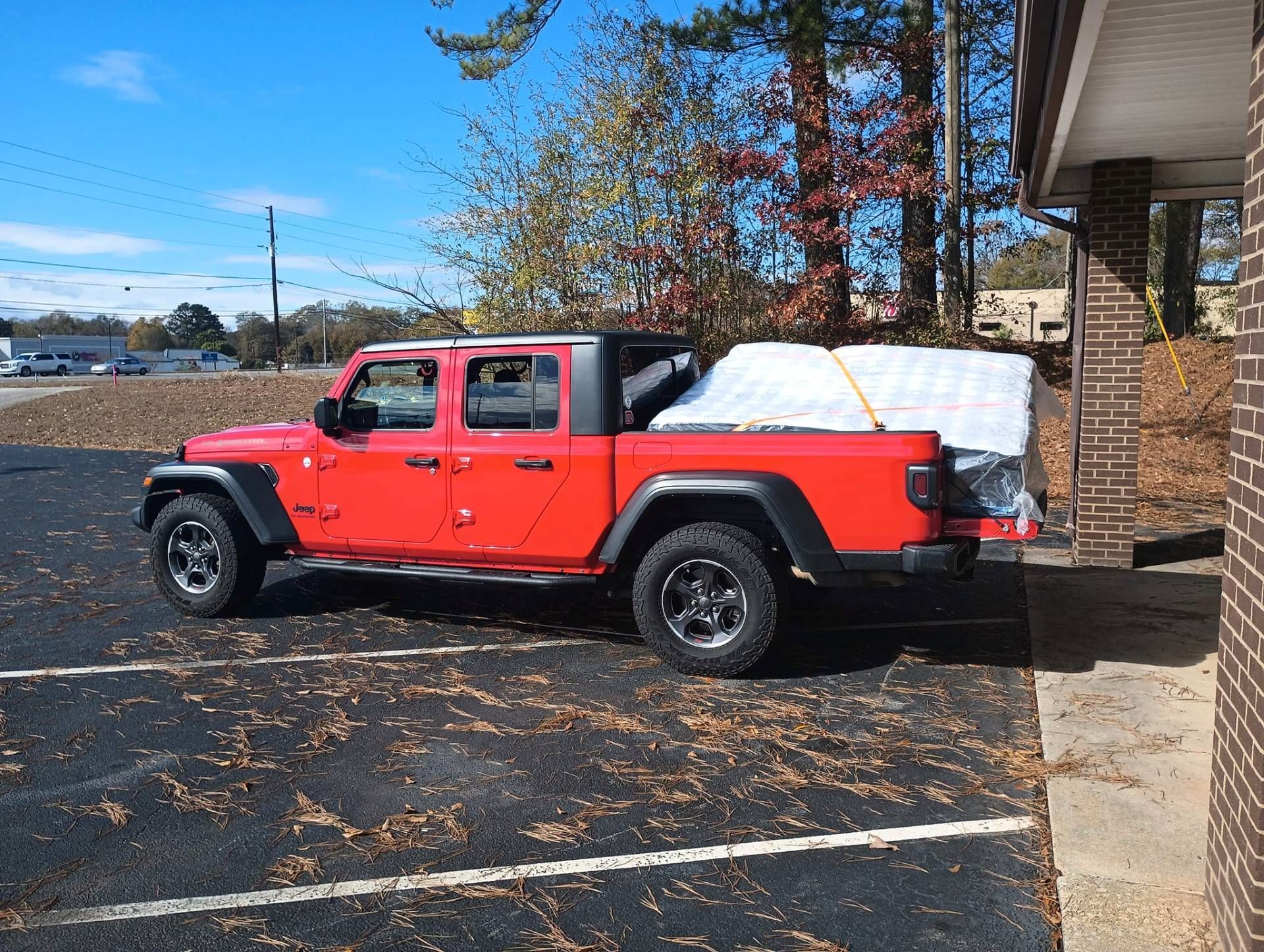 Red Jeep Gladiator pickup truck with a mattress in the bed, parked outside a building.