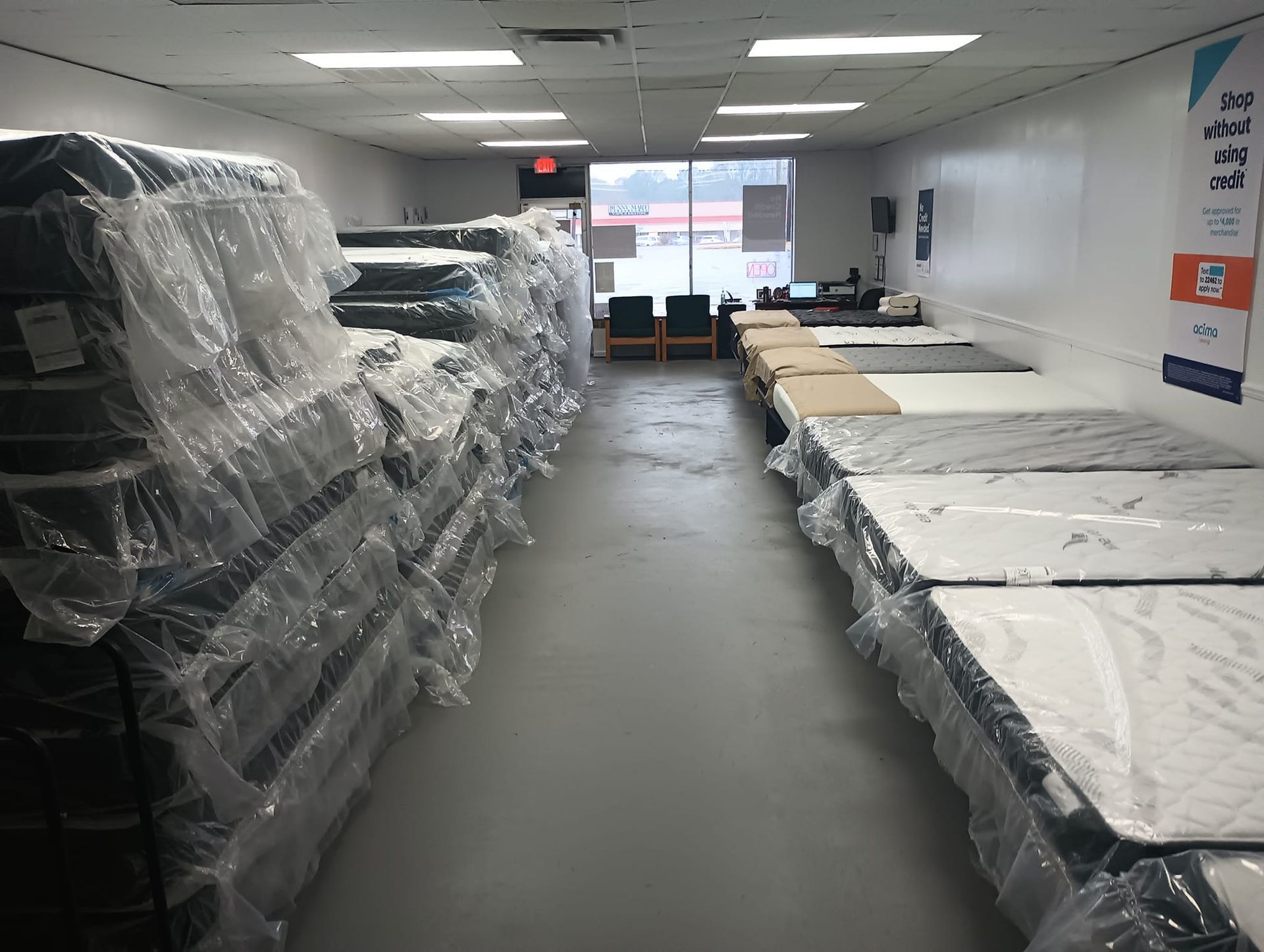 Mattress showroom with stacks of wrapped mattresses and display models on a gray floor.