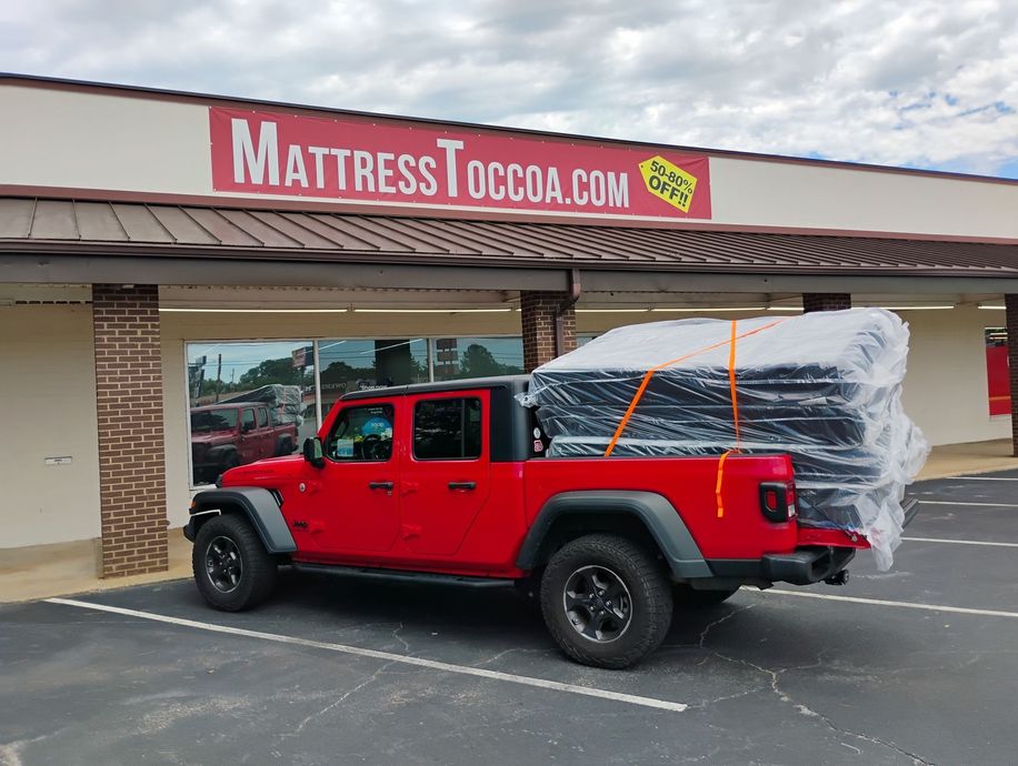 Red Jeep pickup truck with mattresses strapped in the back, parked outside MattressToccoa.com store.