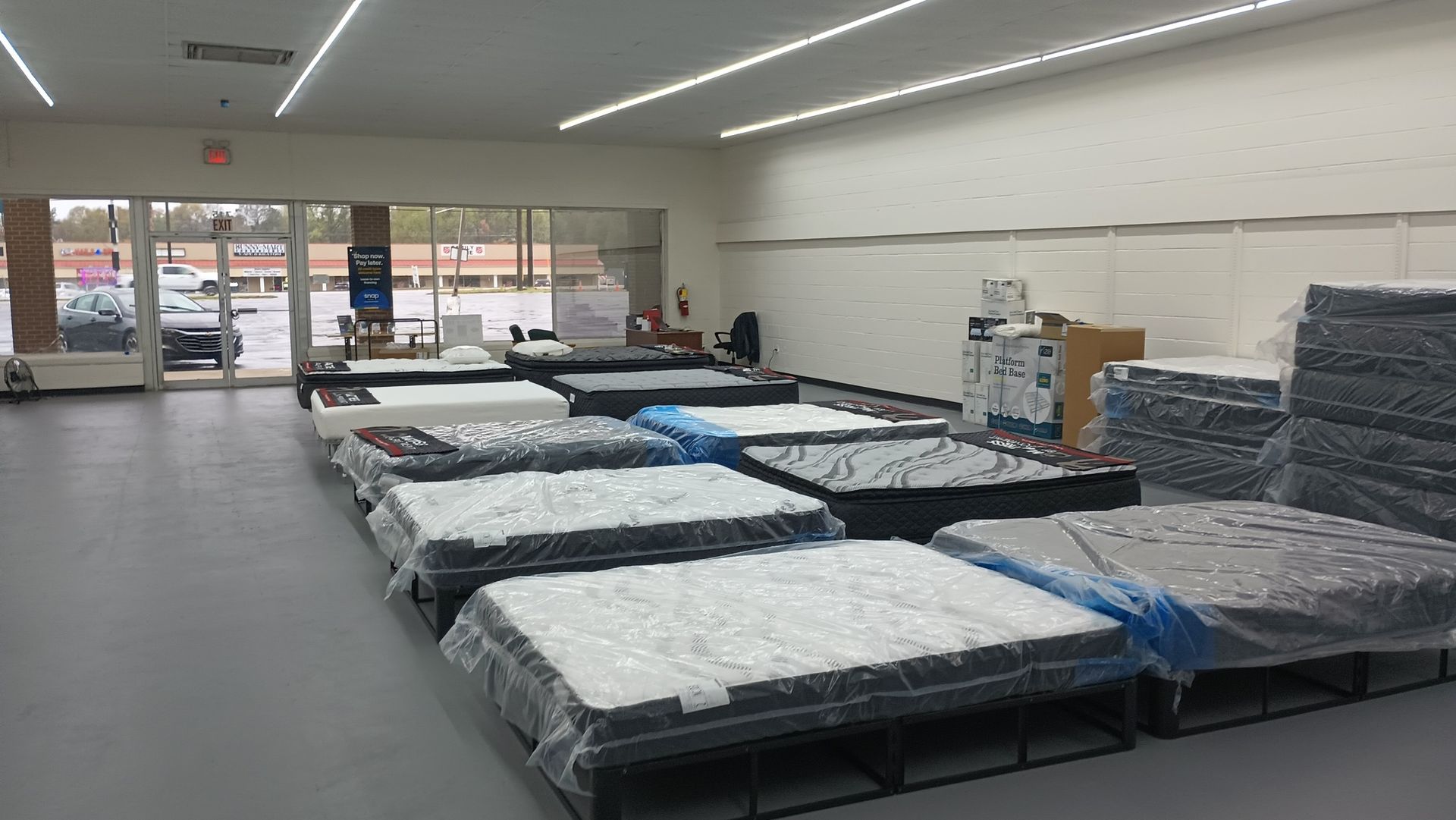 Mattress store interior with rows of beds covered in plastic wrap. Bright lighting, view of outside through glass door.