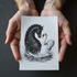 Hands holding a drawing of a black horse and white swan in water.