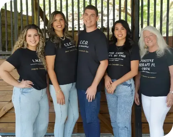 Five people standing together outdoors, wearing matching dark shirts and jeans in front of a wooden fence.