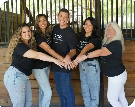 Five people in black shirts and jeans stand together, stacking hands and smiling outdoors.