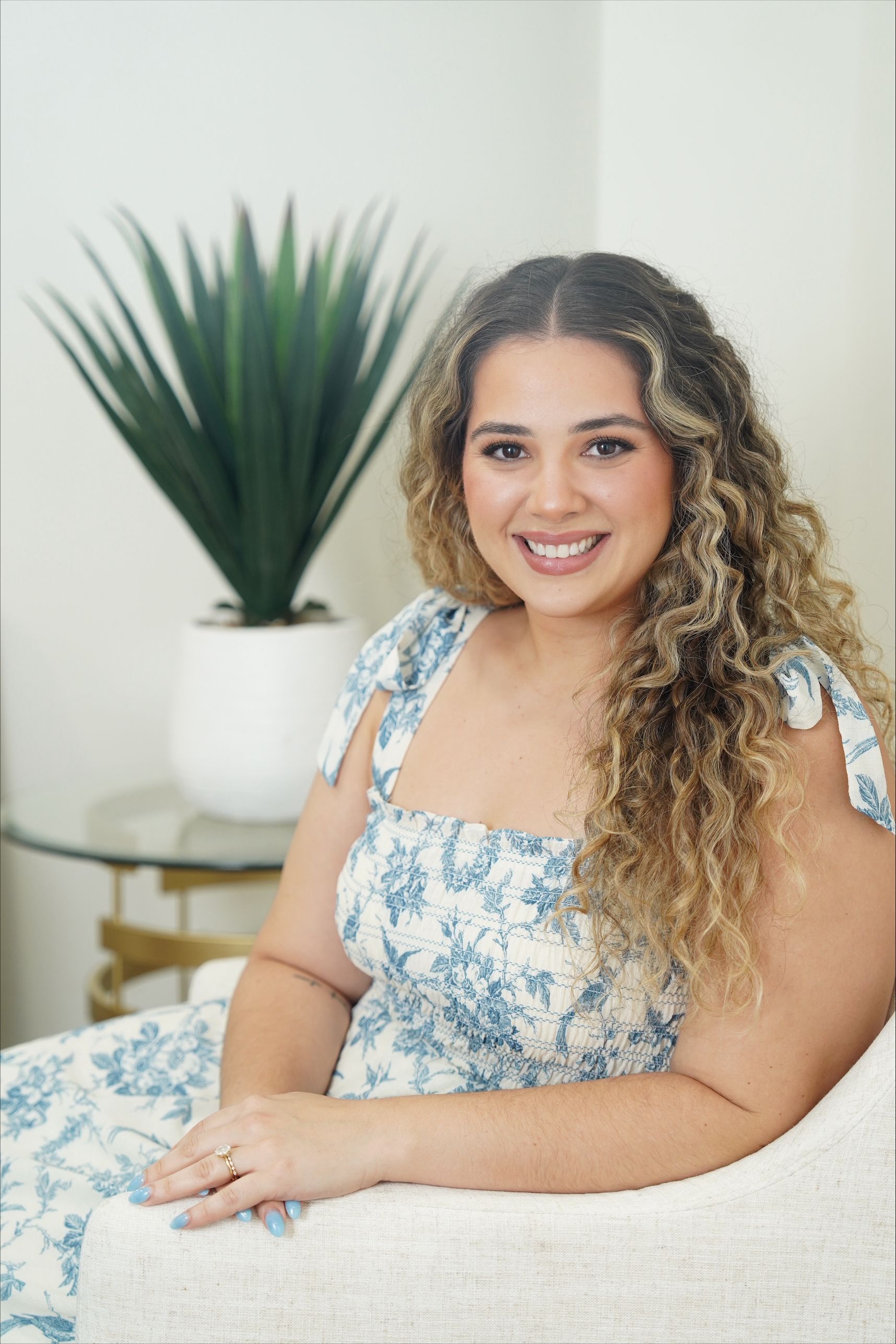 Smiling woman in a blue floral dress seated on a white couch beside a potted plant