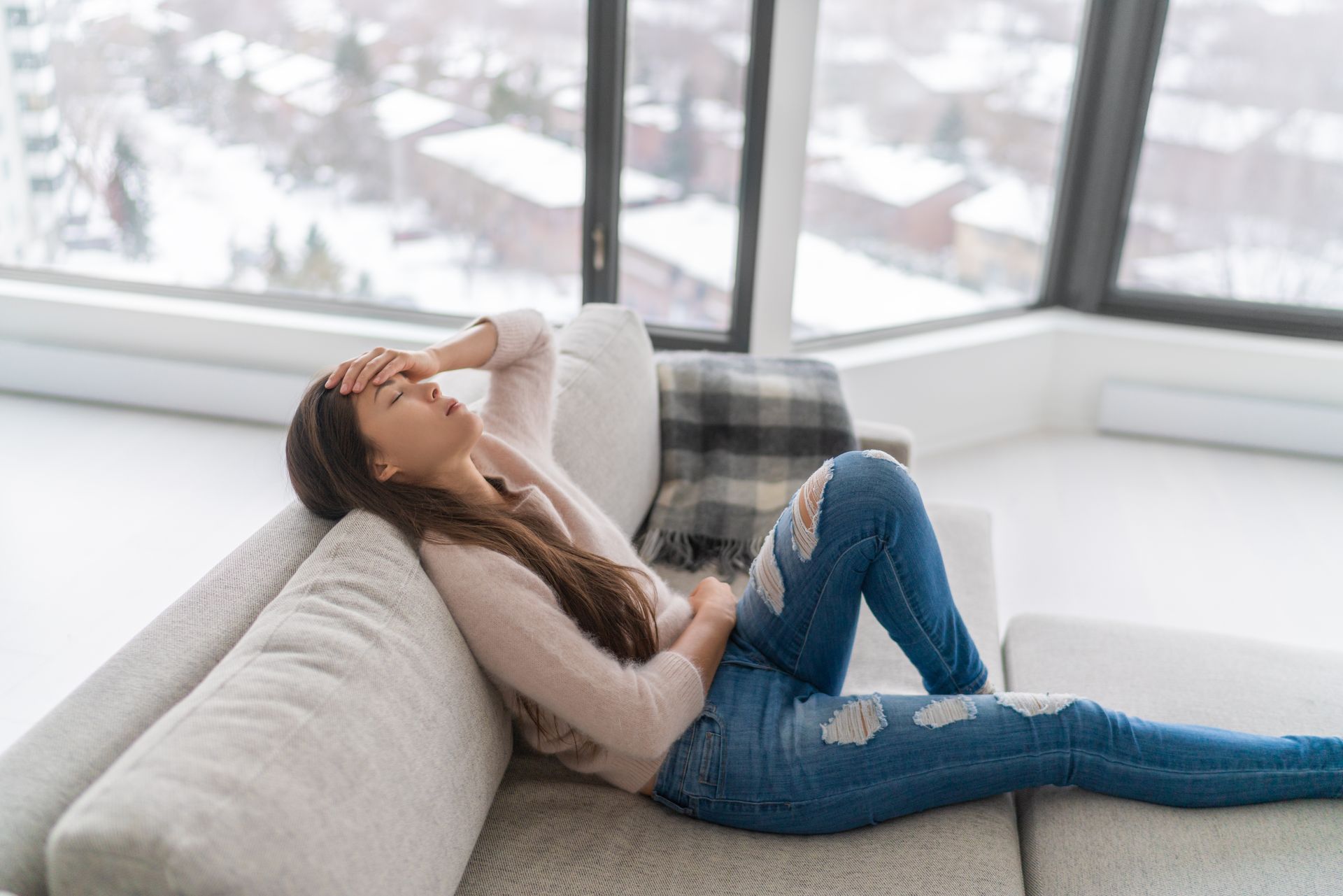 Woman reclining on a gray sofa by a window, wearing ripped jeans and a white sweater.