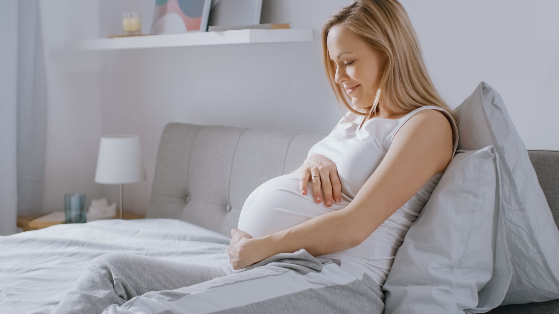 Pregnant woman sitting on a bed, gently holding her belly in a bright bedroom.