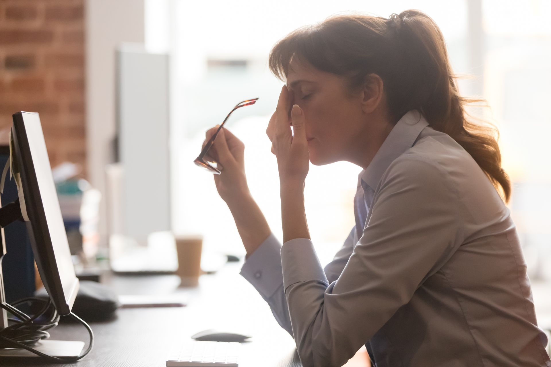 Person at a desk rubbing their eyes while holding glasses, with a computer monitor nearby and bright backlight