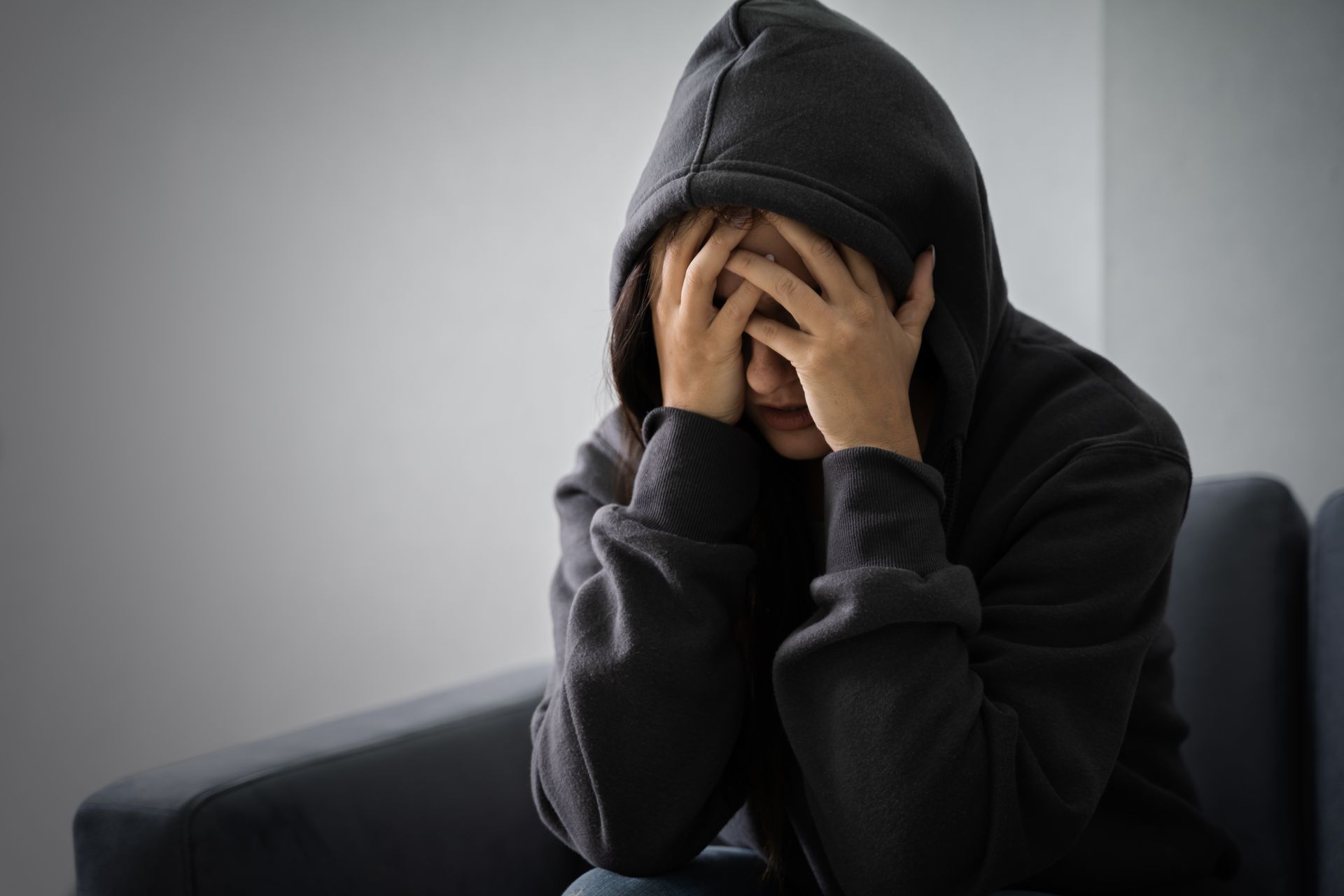 Person in a dark hoodie sits on a couch with hands covering their face, looking distressed.