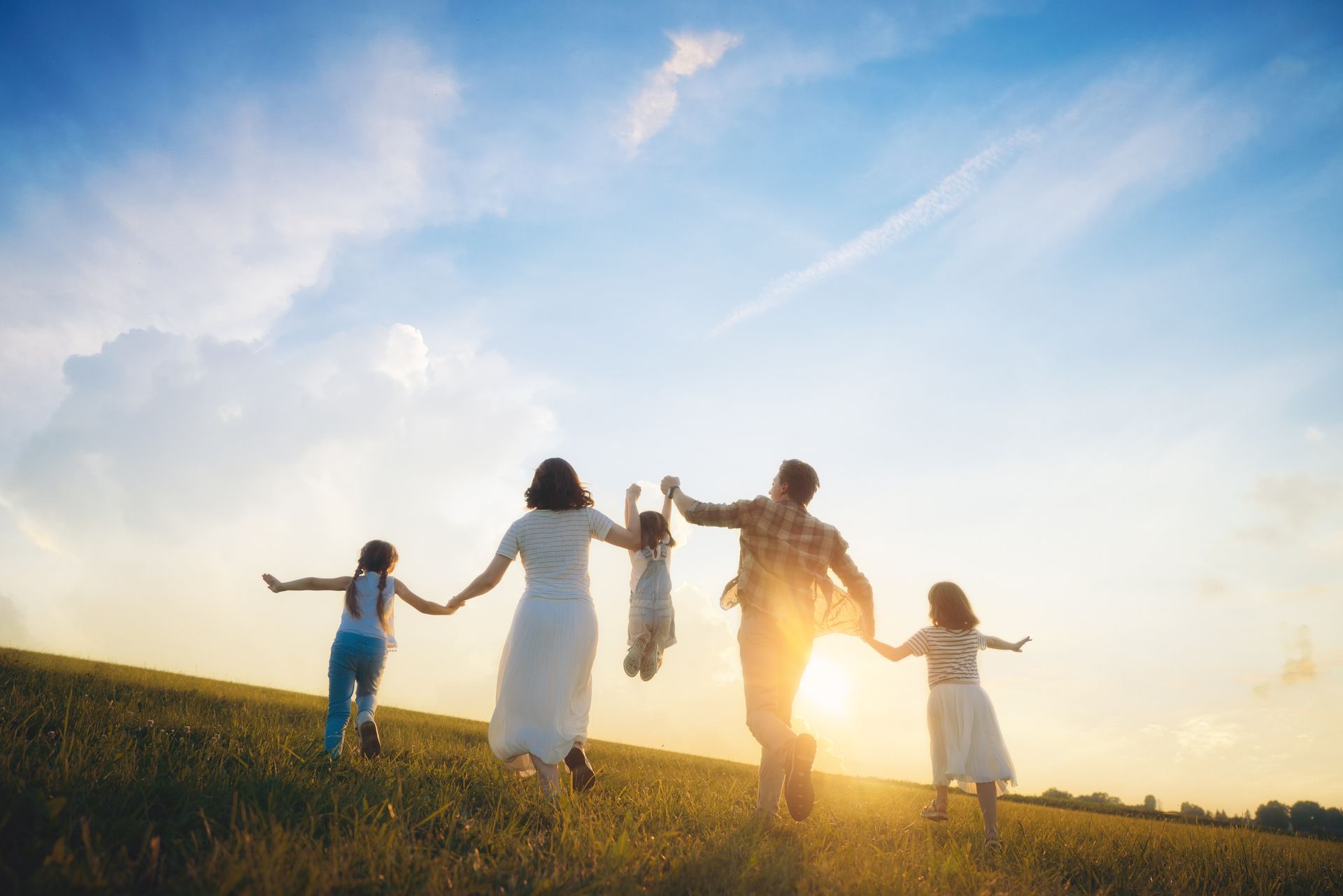 Family holding hands in a grassy field at sunset, backlit by a warm golden sky